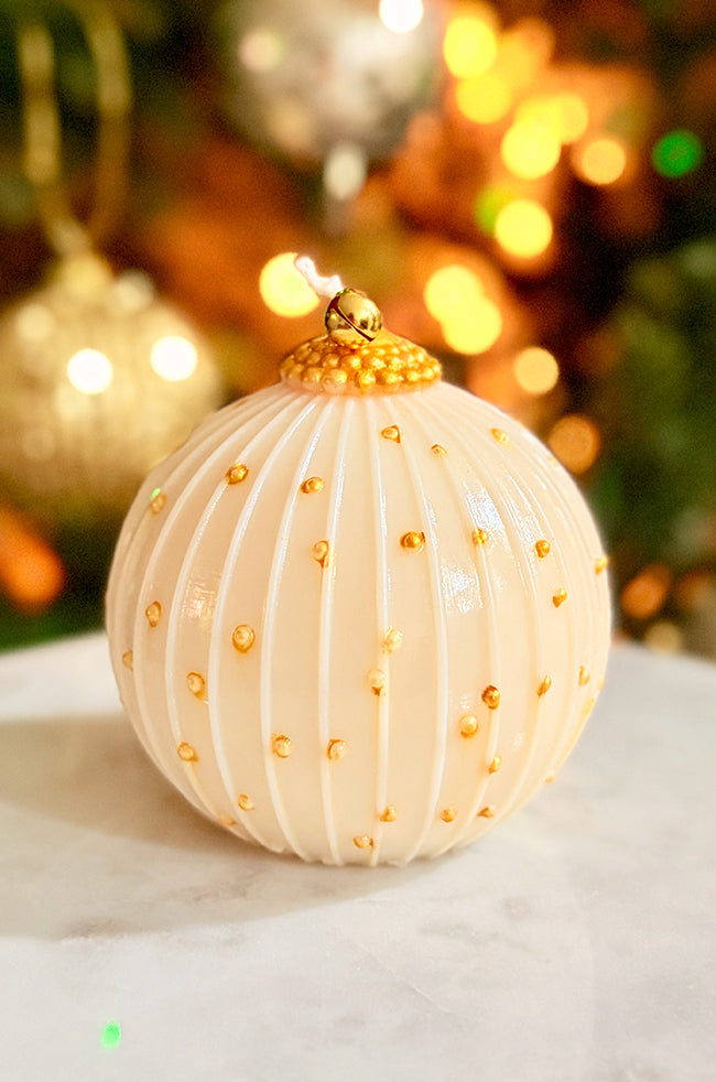 Golden Royale Ornament Candle: Hand-poured ivory ornament candle with vertical ridges, gold hand-painted accents, and a golden bell topper, displayed on a marble surface with warm holiday lights in the background.