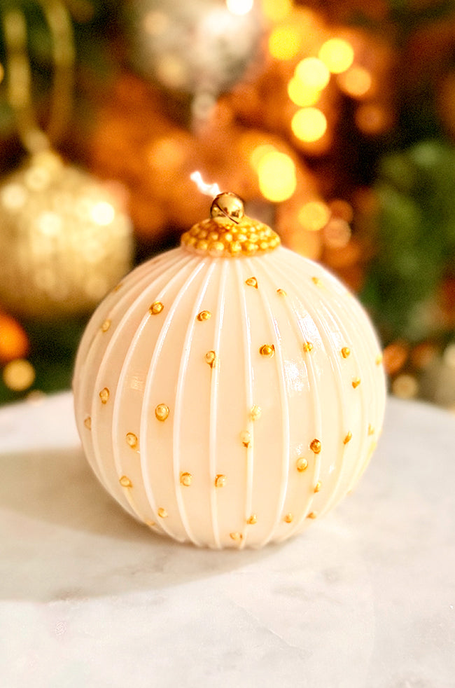 Golden Royale Ornament Candle: Close-up of the Golden Royale Ornament candle showing its glossy varnish finish, gold dotted accents, and bell topper against festive, glowing holiday lights.