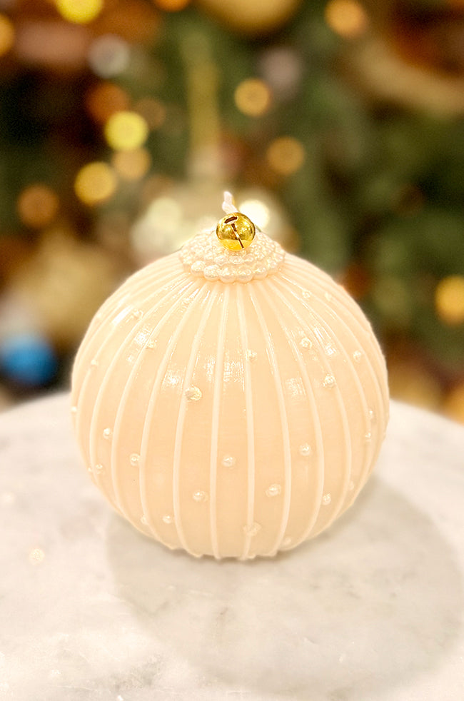 Golden Royale Ornament Candle: Ornament candle with glossy ribbed texture and delicate pearl accents, topped with a gold bell, placed on a marble surface with festive warm lighting behind it.