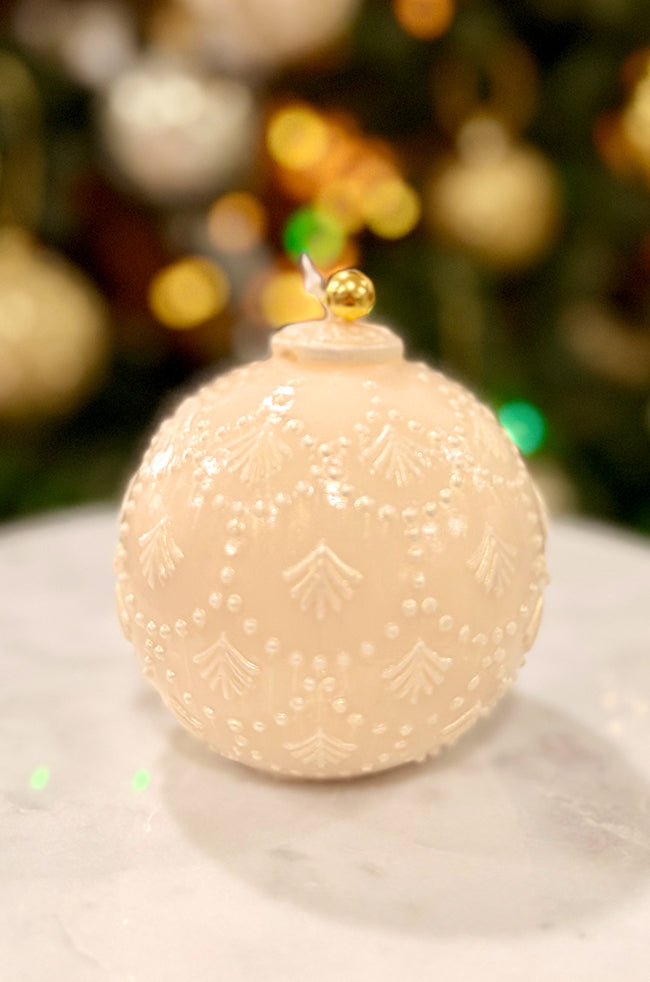 Golden Star Ornament Pear Candle: Ivory pearl-toned ornament-shaped candle with raised dotted and leaf-pattern detailing, topped with a small gold bell, photographed on a marble surface with warm holiday lights blurred in the background.