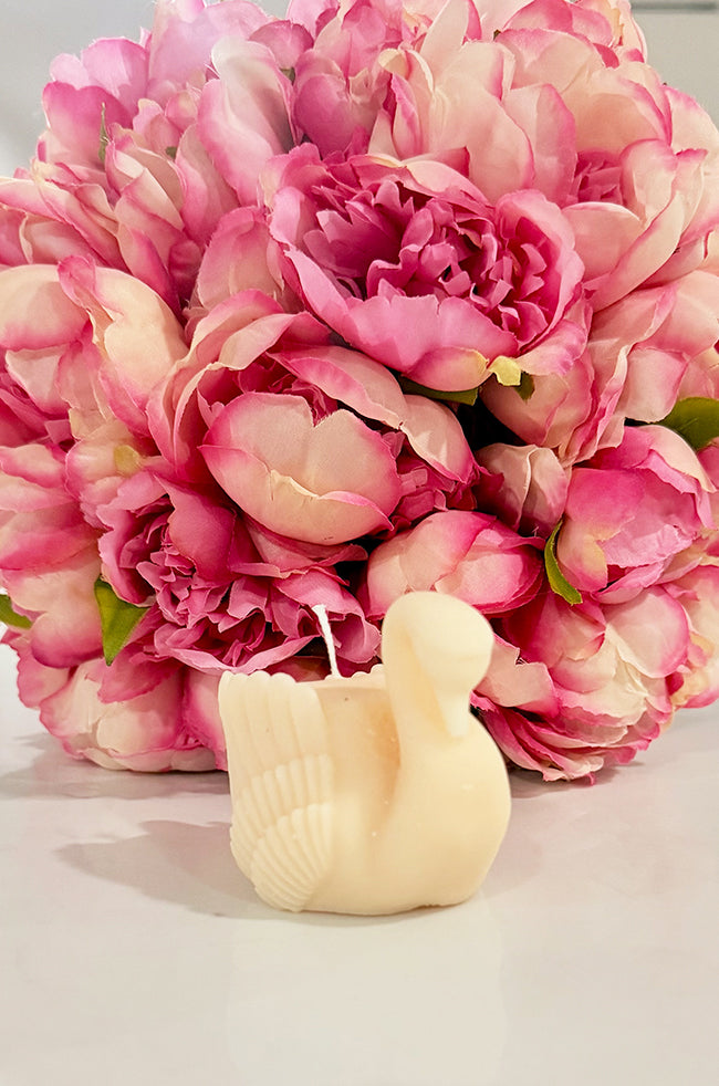 Grace Swan Candle: Handcrafted ivory Grace Swan sculptural candle displayed in front of a bouquet of pink peonies, showcasing its elegant swan silhouette and detailed feather design.