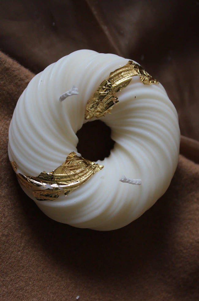 Gyrate Girdle Candle: Ivory round sculptural candle with grooved texture and gold foil accents, displayed on a brown fabric background.