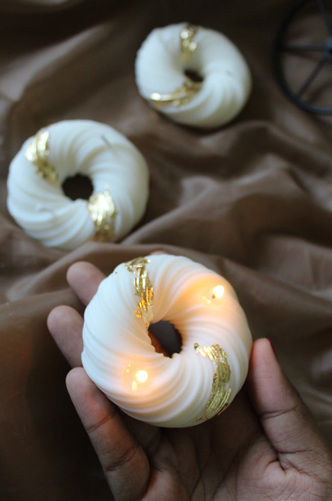 Gyrate Girdle Candle: Hand holding a lit ivory sculptural candle with gold foil accents, with additional gold-accented round candles displayed on brown fabric in the background.