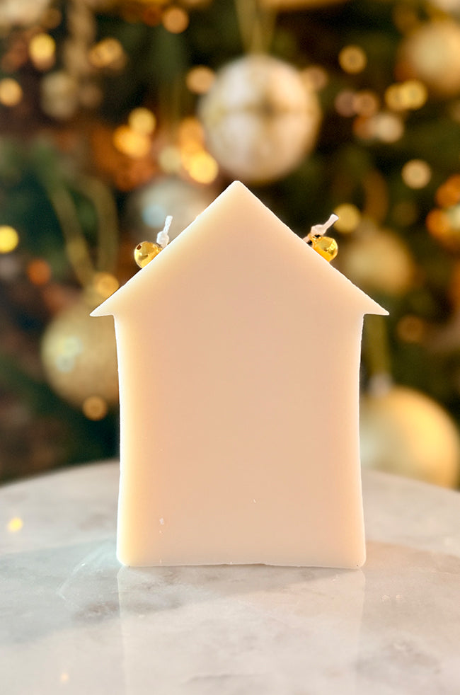 Home for Christmas Candle: Back view of the ivory Home for Christmas house-shaped candle with two golden bells and a softly lit Christmas tree background.