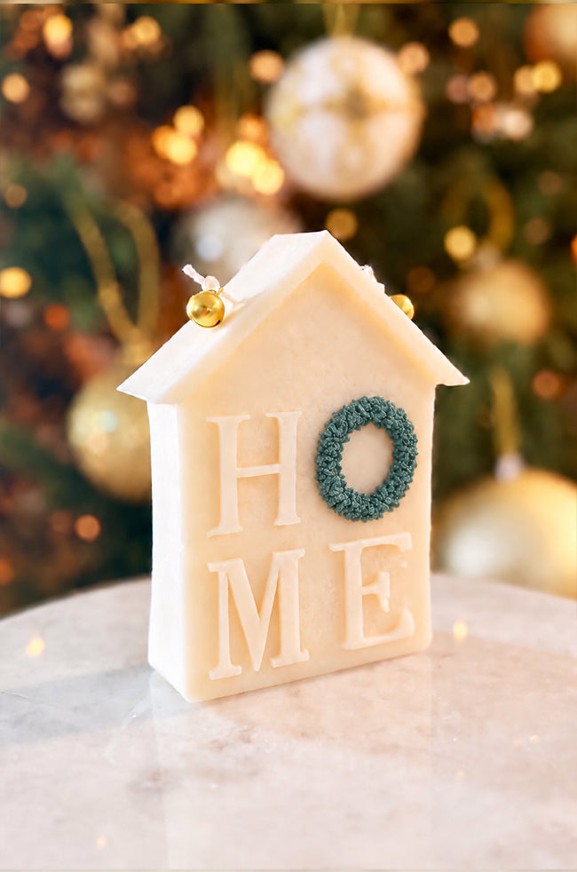 Home for Christmas Candle: Angled view of the Home for Christmas sculptural candle featuring HOME lettering, with a green wreath detail and two golden bells against festive holiday décor.