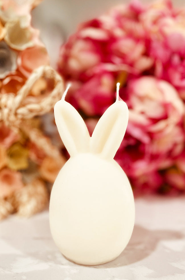Hop Bunny Candle: White bunny-shaped candle with tall ears, placed in front of pink flowers.