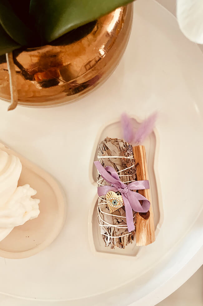Healing Energy Enhancer Lavender Heart Evil Eye Charm Kit: Lavender, sage, and Palo Santo bundle tied with a purple ribbon and adorned with an Evil Eye charm, displayed on a white tray.