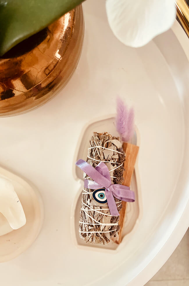 Healing Energy Enhancer Lavender Oval Evil Eye Charm Kit: Lavender, sage, and Palo Santo bundle tied with a purple ribbon and adorned with an Evil Eye charm, displayed on a white tray.