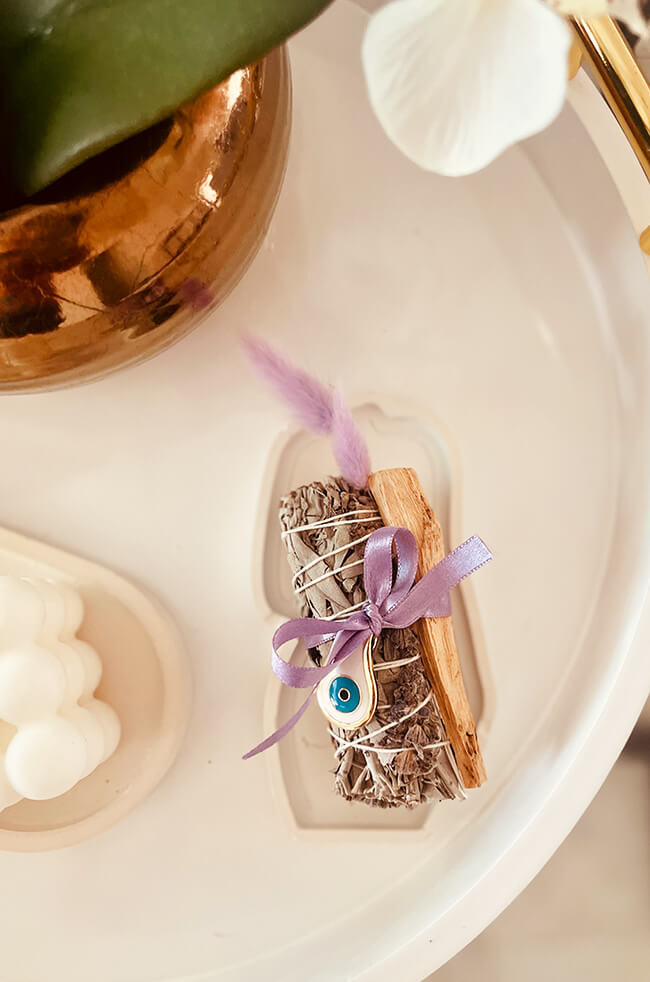Healing Energy Enhancer Lavender Rain Drop Evil Eye Charm Kit: Lavender, sage, and Palo Santo bundle tied with a purple ribbon and adorned with an Evil Eye charm, displayed on a white tray.