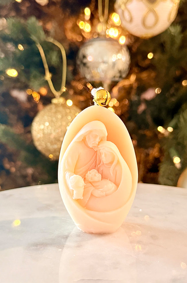 Nativity Glow Candle: Ivory Nativity Glow sculptural candle featuring the Holy Family and a gold bell, displayed in front of a decorated Christmas tree.