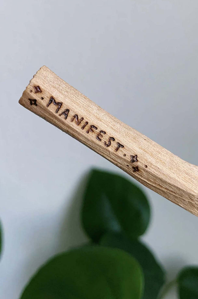 Palo Santo: Single Palo Santo stick engraved with the word “Manifest,” photographed against a neutral background with green leaves.