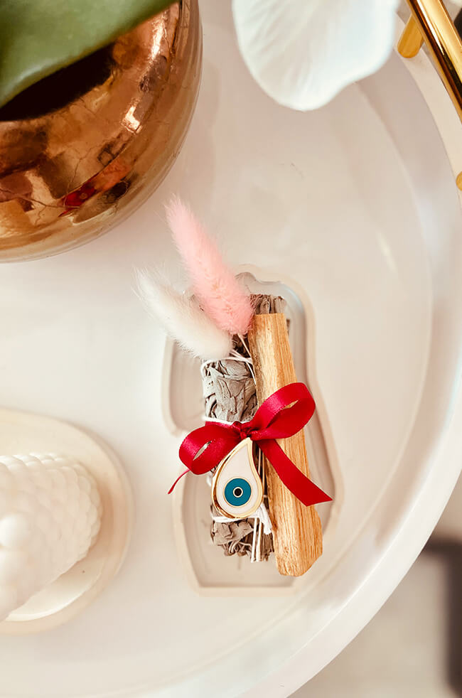 Healing Energy Enhancer White Sage Rain Drop Evil Eye Charm Kit: White sage and Palo Santo bundle tied with a red ribbon and adorned with an Evil Eye charm, displayed on a white tray.