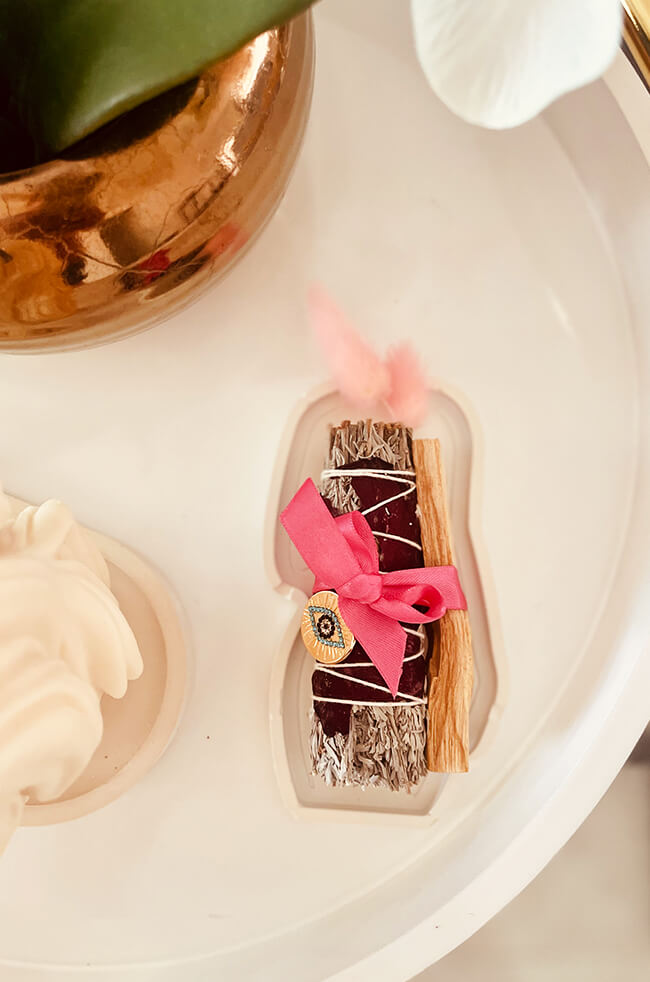 Healing Energy Enhancer Rose Circle Evil Eye Charm Kit: Rose, sage, and Palo Santo bundle tied with a pink ribbon and adorned with an Evil Eye charm, displayed on a white tray.
