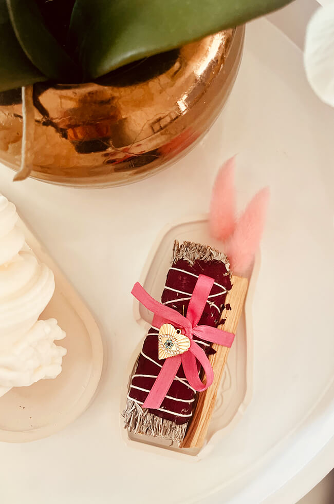 Healing Energy Enhancer Rose Heart Evil Eye Charm Kit: Rose, sage, and Palo Santo bundle tied with a pink ribbon and adorned with an Evil Eye charm, displayed on a white tray.