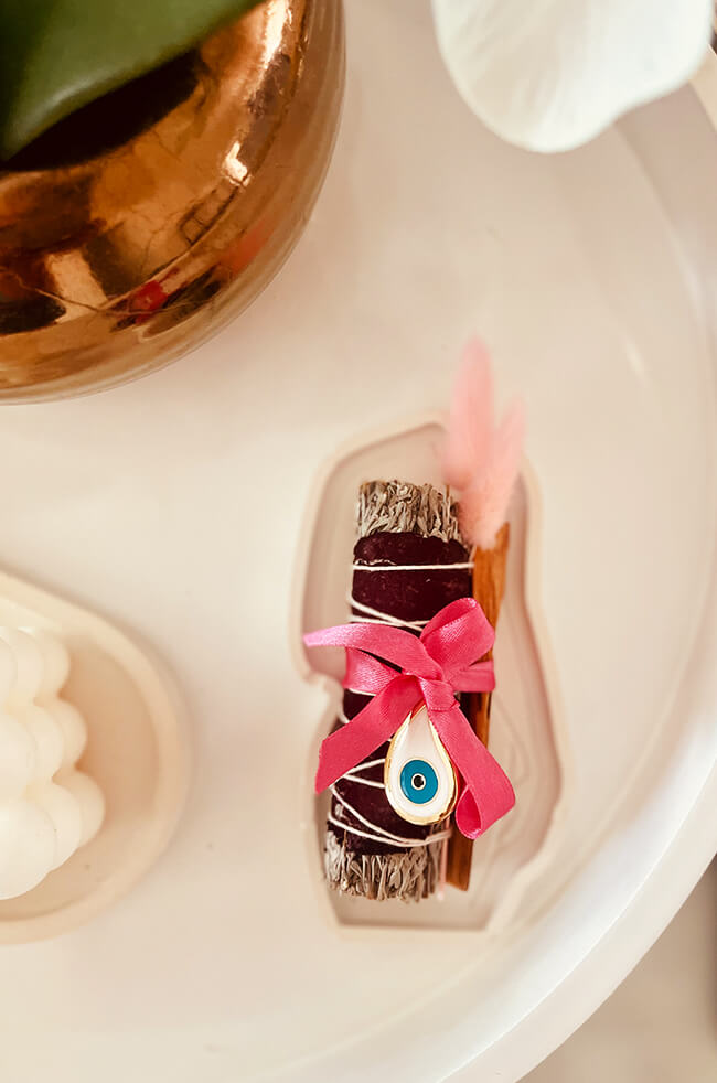 Healing Energy Enhancer Rose Rain Drop Evil Eye Charm Kit: Rose, sage, and Palo Santo bundle tied with a pink ribbon and adorned with an Evil Eye charm, displayed on a white tray.
