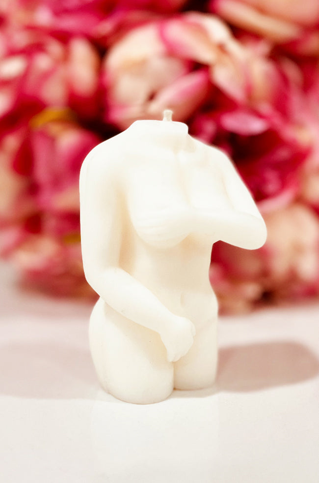 Shy Lady Candle: Angled view of the Shy Lady sculptural candle with one arm across the chest and soft floral décor behind it.