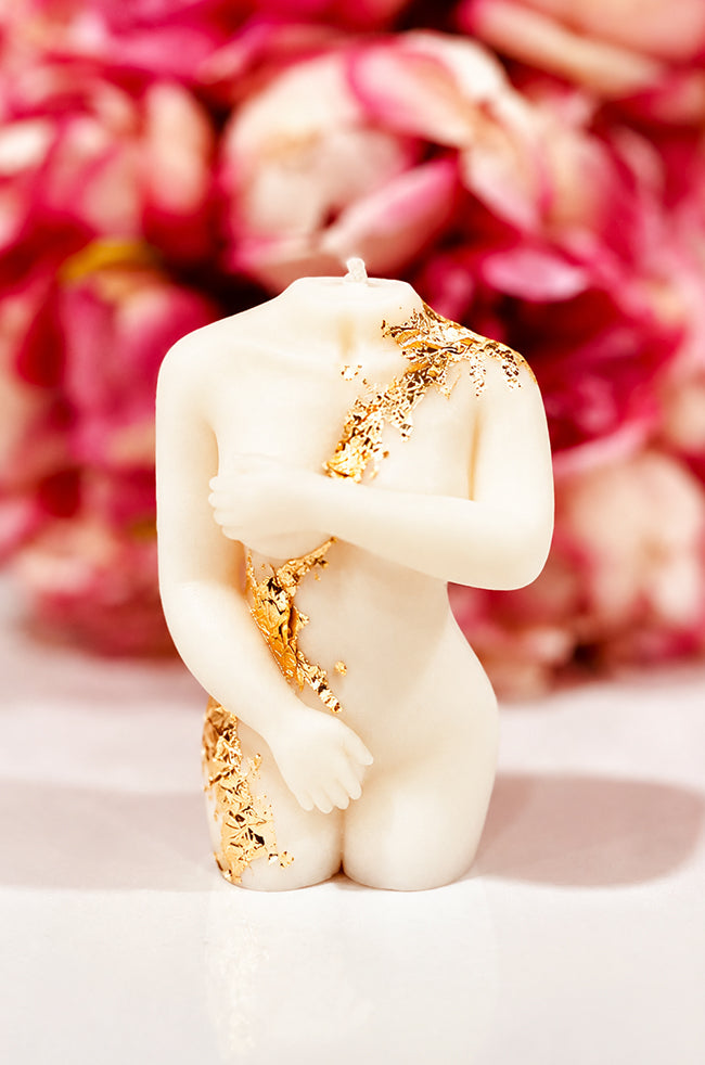 Shy Lady Candle: Shy Lady ivory sculptural body candle with elegant gold leaf accents and a soft pink floral background.