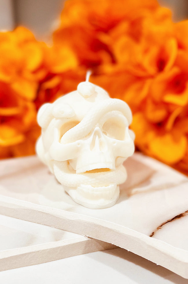 Skull Snake Candle: Front view of the ivory Skull Snake sculptural candle with a snake winding through the skull, styled with orange Halloween flowers.