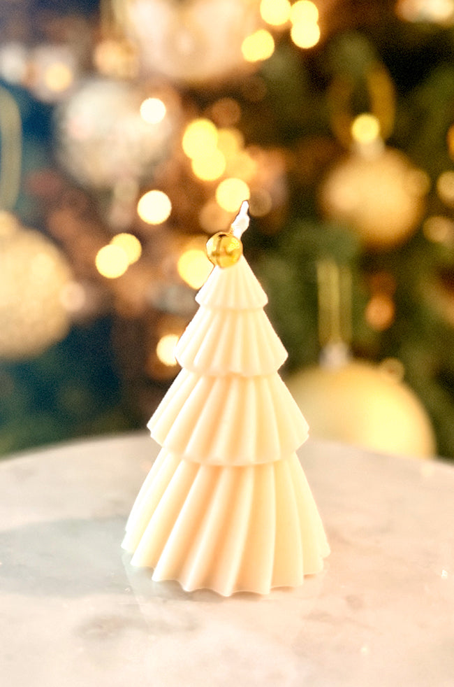 Solstice Tree Small: A fluted cream tree-shaped candle with cascading gold leaf details, topped with a golden bell, sitting on a marble surface with blurred holiday lights in the background.