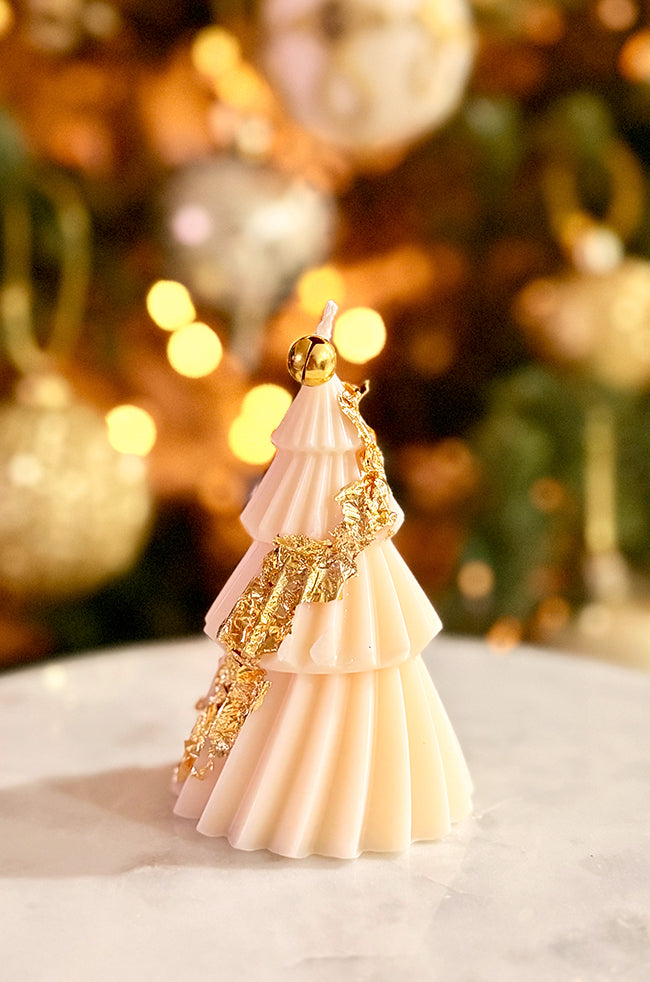 Solstice Tree Small: Festive tree-shaped candle with golden leaf garland details, artfully hand-applied to highlight the folds, glowing in front of a blurred Christmas background.