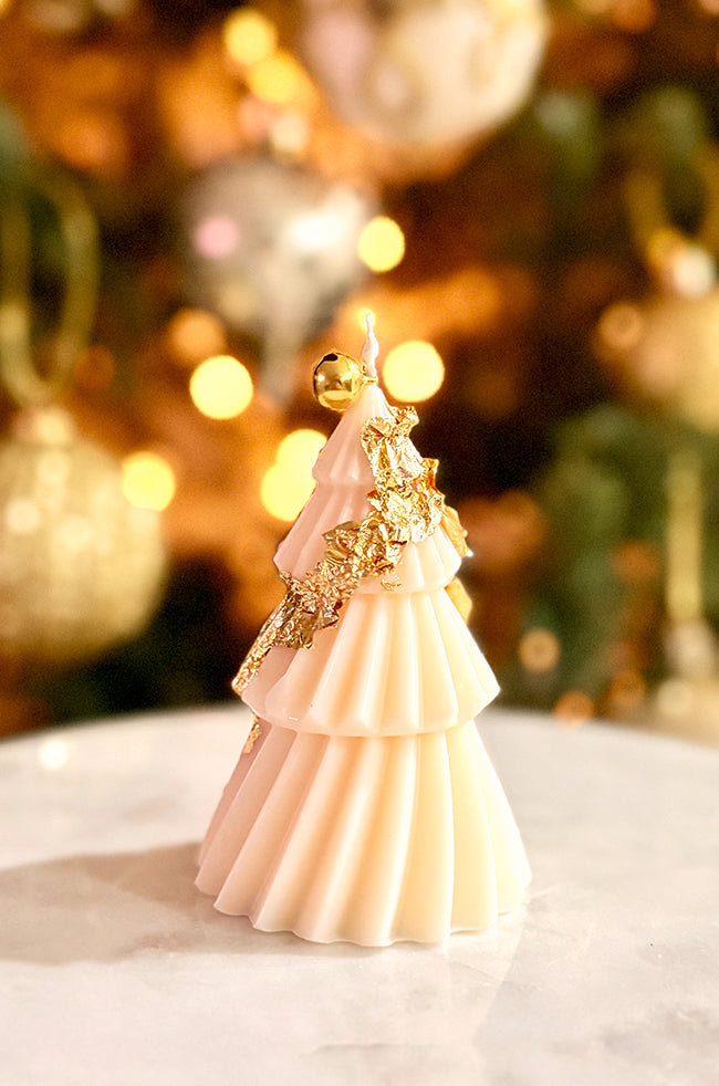 Solstice Tree Small: Minimalist sculpted Christmas tree candle in cream color, standing tall with a golden bell topper, styled with warm fairy lights and ornaments in the background.