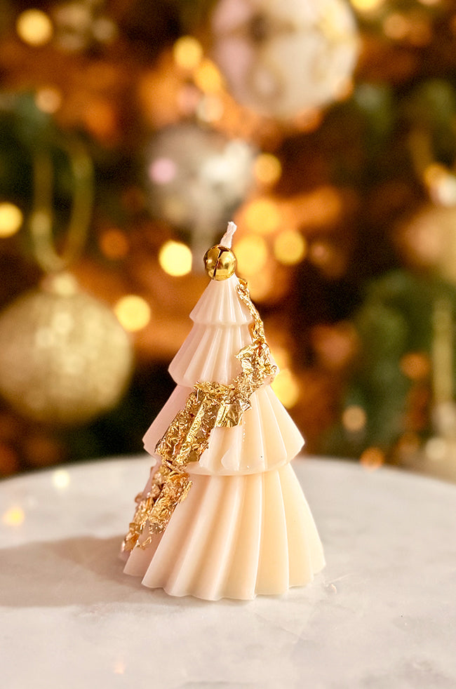 Solstice Tree Small: Small cream-toned tree candle with fluted tiers and a shiny gold bell accent, styled on a marble surface with soft golden holiday lights in the background.