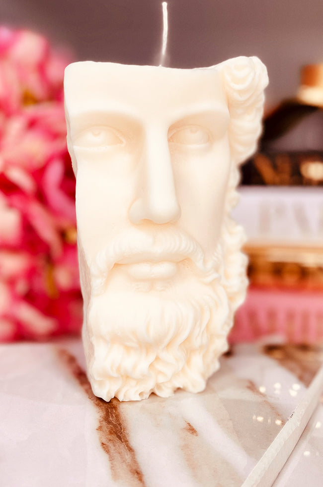 Stoic Candle: Front view of the ivory Stoic sculptural candle with a classical bearded face, styled on a marble surface with pink flowers.
