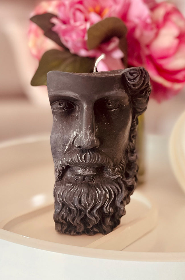 Stoic Candle: Dark charcoal Stoic sculptural candle with detailed facial features and beard, displayed with pink floral décor.