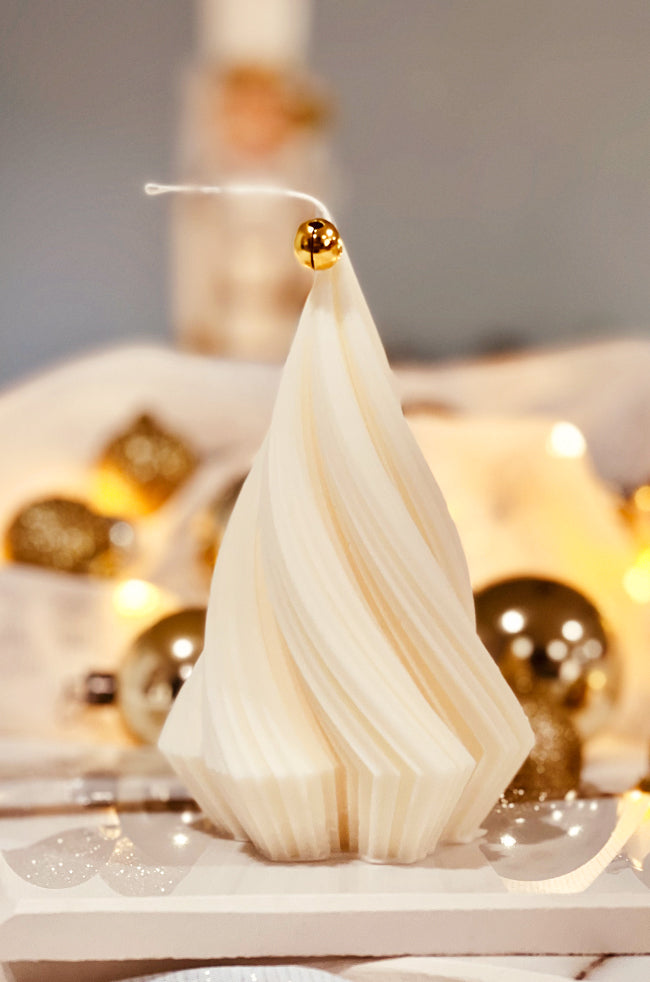 Swirling Fir Tree Large Candle: A sculpted white candle shaped like a modern Christmas tree with swirling folds, topped with a gold bell, surrounded by golden holiday ornaments and soft festive lighting.