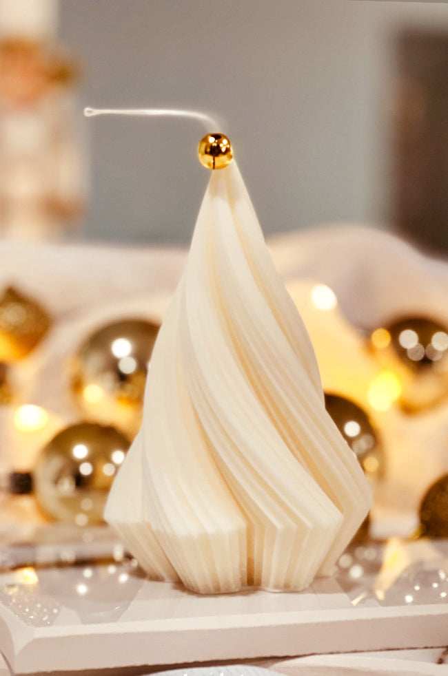 Swirling Fir Tree Large Candle: Hand-poured white sculptural candle resembling a spiraled fir tree, featuring a golden bell on the wick, staged with reflective gold ornaments and a cozy holiday ambiance.