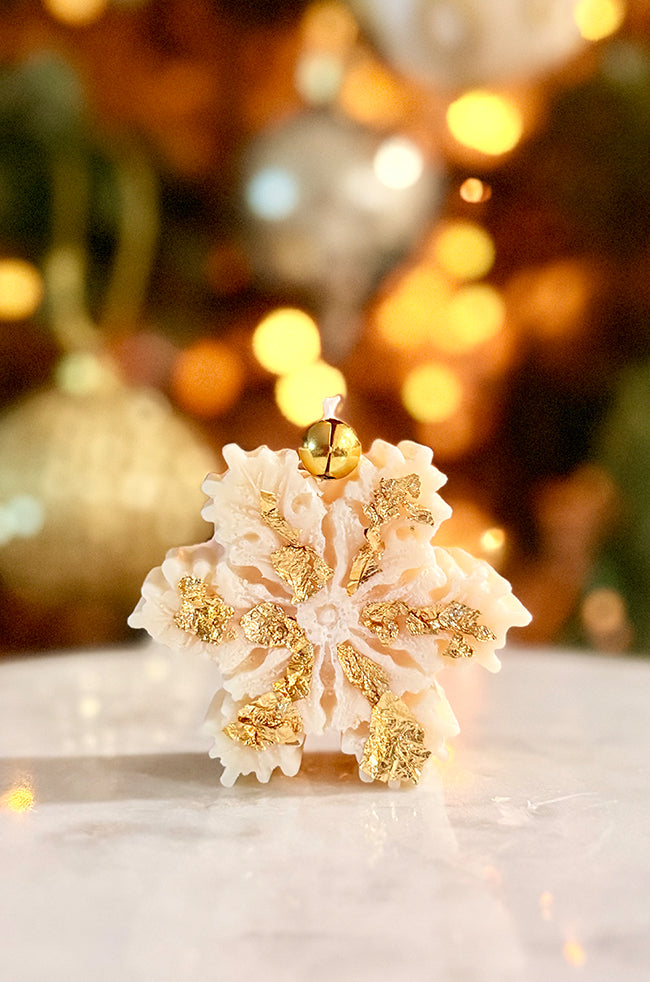 The Christmas Snowflake Candle: Front view of The Christmas Snowflake sculptural candle with gold flakes and gold bell against festive holiday lights.