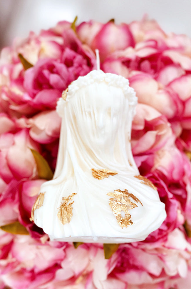 The Veiled Maiden Candle: Luxury handmade Veiled Maiden candle with gold leaf accents, inspired by classical sculpture, displayed on pink floral background.